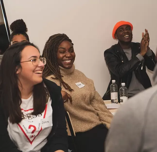 A group of people are sitting and smiling in a well-lit room. One person is clapping, wearing an orange hat and black jacket. Others are wearing casual clothing and name tags. Water bottles are placed on the tables in front of them by Quintessentially Foundation.