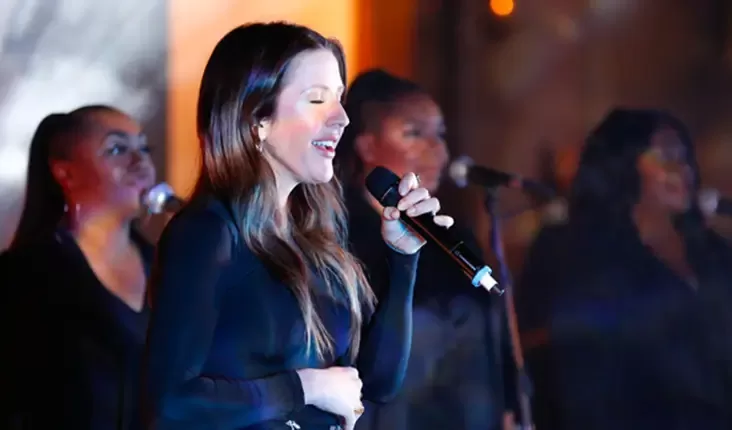 Ellie Goulding, the famous singer, is singing into a microphone on stage, with three backup singers in the background. They are all dressed in black, and the stage lighting creates a warm, ambient atmosphere at Quintessentially Foundation's Fayre of St James annual concert.
