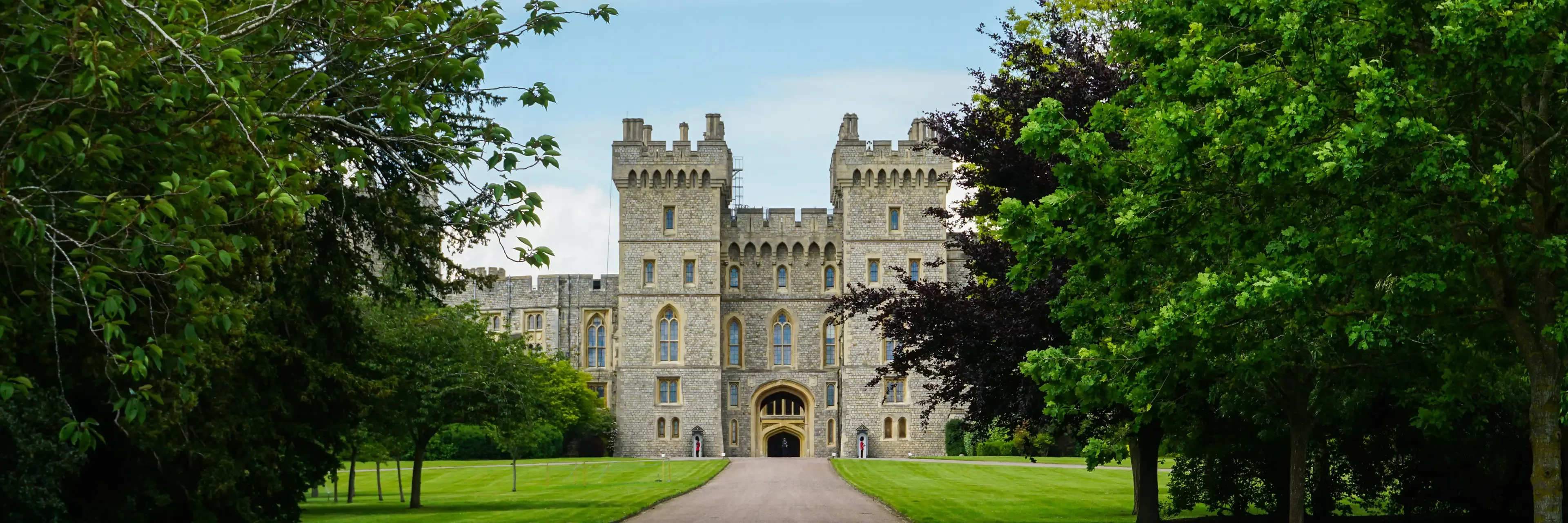 A grand stone castle with tall towers and crenellated walls is seen at the end of a tree-lined pathway, surrounded by lush green lawns and leafy trees under a partly cloudy sky.