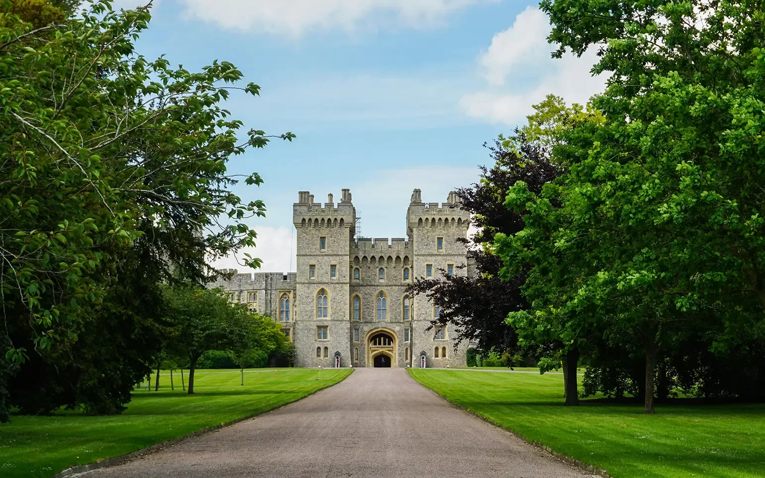 A long gravel path leads up to a large stone castle with battlements, arched windows, and an ornate entrance, surrounded by lush green lawns and trees under a partly cloudy blue sky.