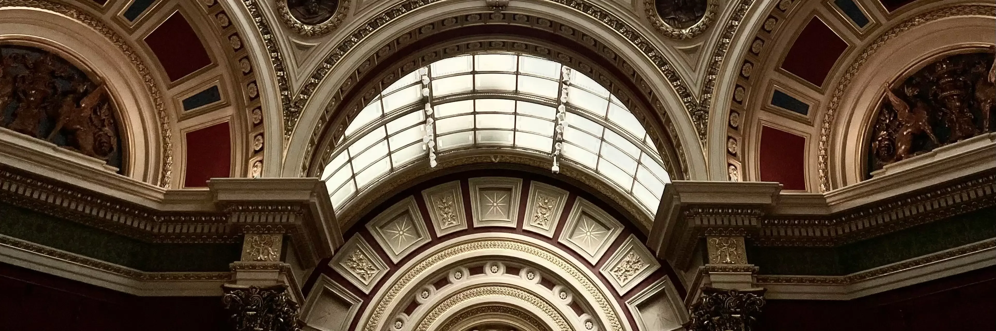 Ornate arched ceiling with intricate carvings, classical columns, and a large skylight. Decorative details in gold, cream, and red create a grand, classical architectural feel.