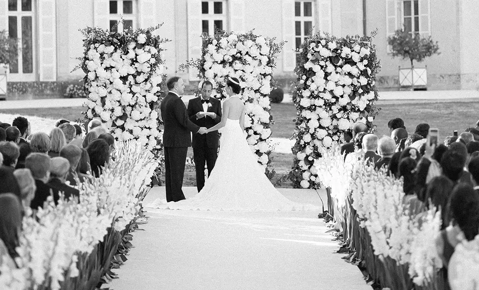 Black and white photo of bride, groom, and officiator down the aisle surrounded by viewers and floral decor outdoors. | Quintessentially Weddings