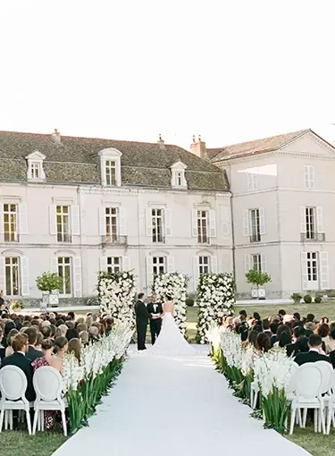 Beautiful white wedding outdoors of an estate with a white aisle, white chairs, and floral decorations with viewers, bride, groom, and officiatorn | Quintessentially Weddings