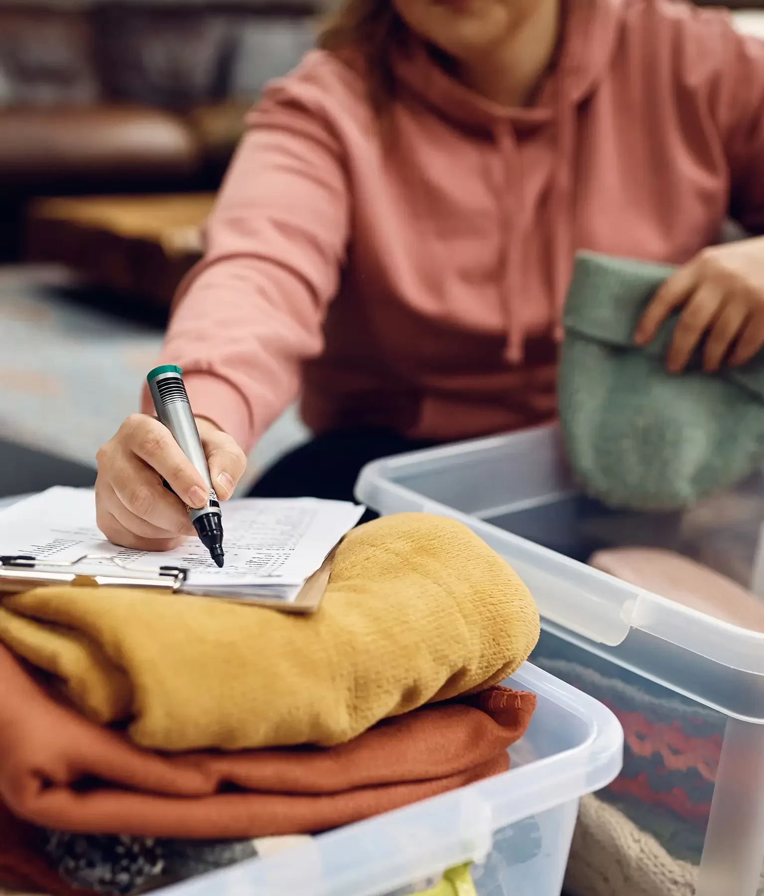 A person in a pink sweatshirt is sitting on the floor, using a black marker to write on a clipboard with papers. They are organizing clothes, including yellow, orange, and green items, into clear plastic bins.