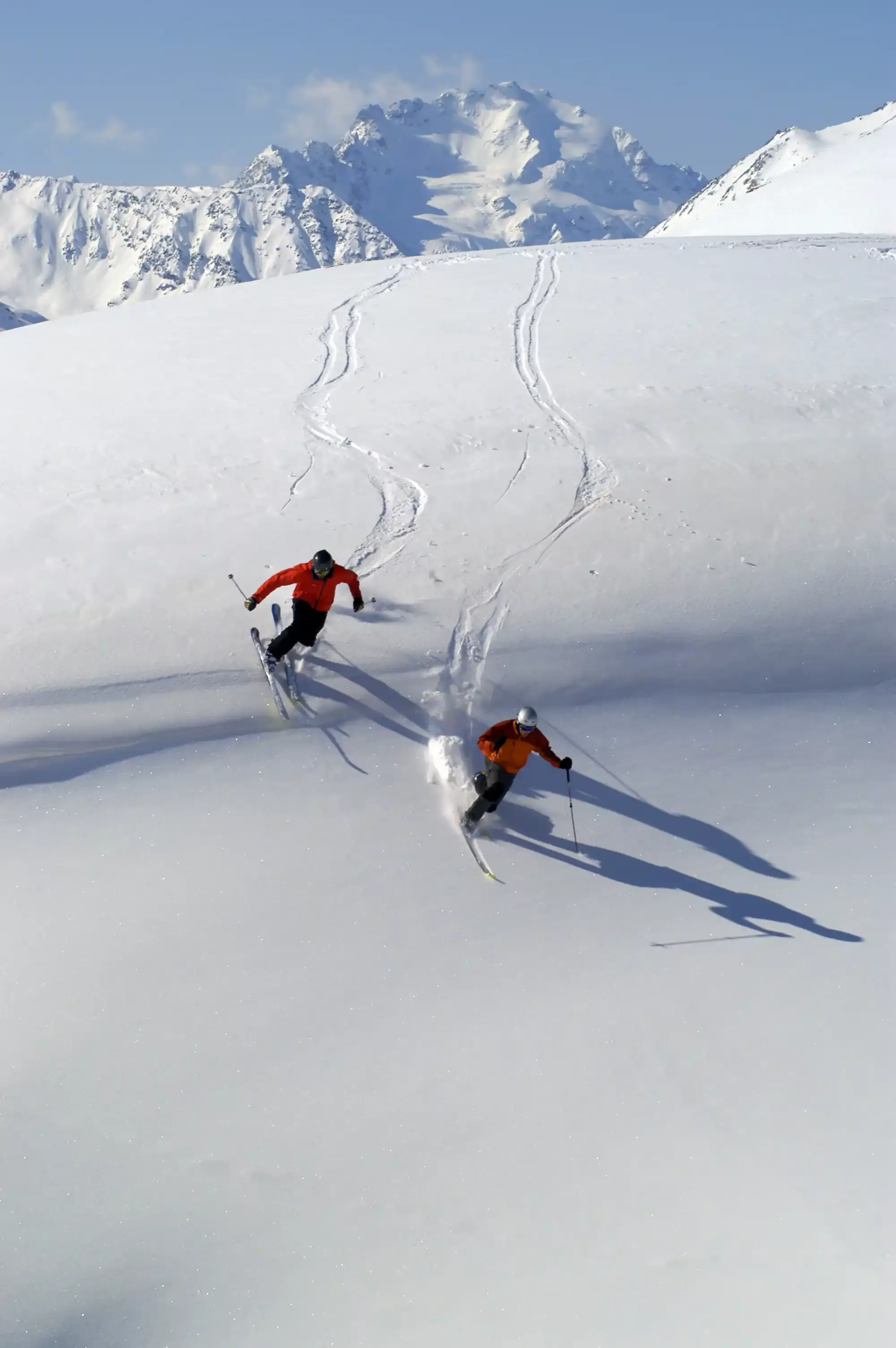 Two people dressed in red ski gear skiing down steep ski slope on mountain.