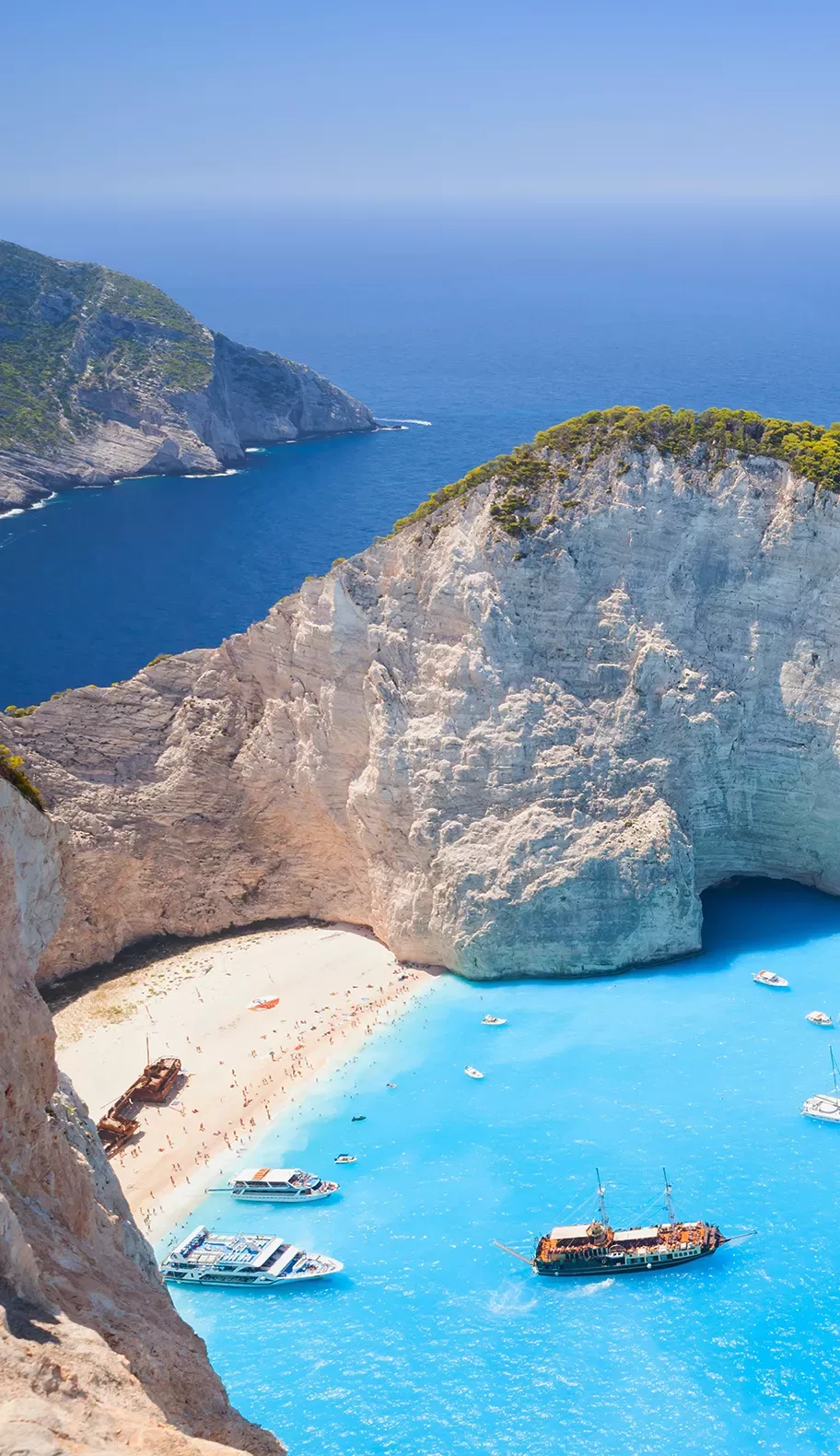 Aerial view of Navagio Beach in Zakynthos, Greece, showing white cliffs, turquoise water, several boats in the bay, and people relaxing on the sandy shore.