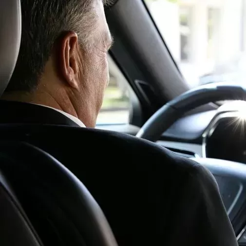 A man in a suit is driving a car, viewed from the back seat. Sunlight is reflecting off the dashboard, and his hands are on the steering wheel as he looks forward.