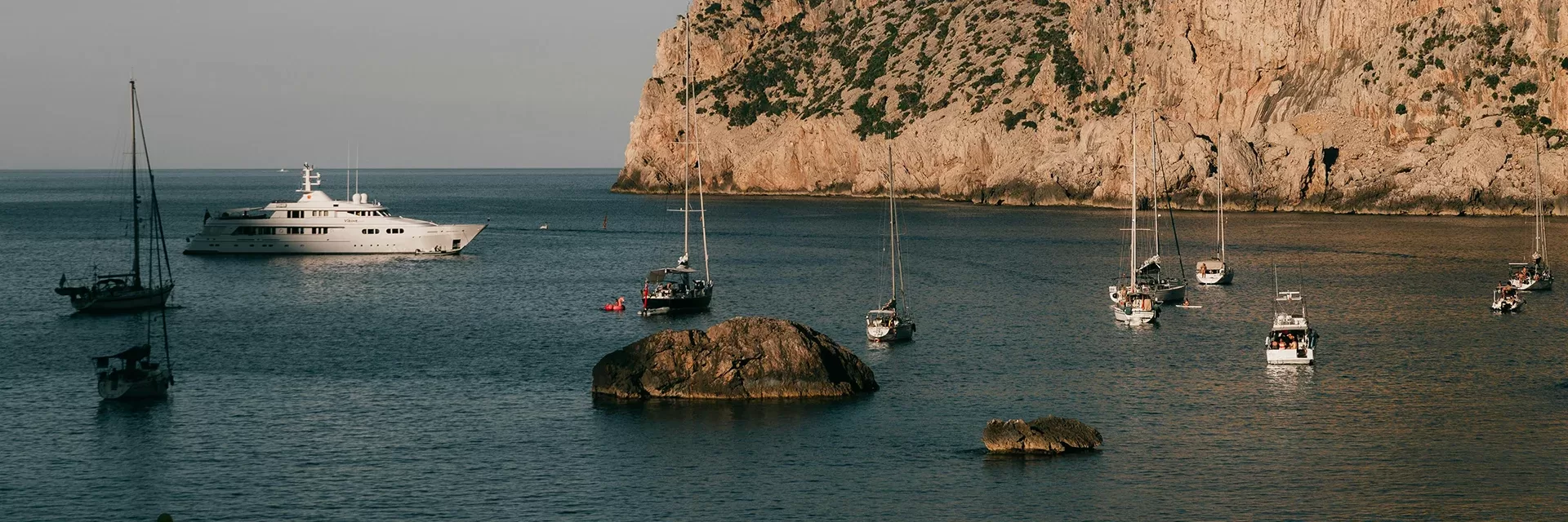Several sailboats and a large yacht are anchored in a calm bay with rocky cliffs and a few large rocks in the water, under clear skies.