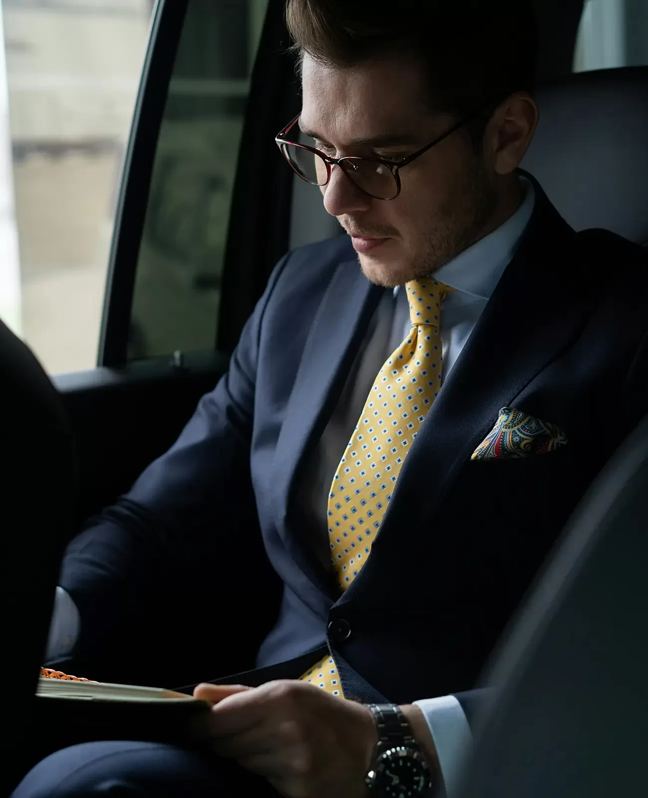 A man in a navy suit, yellow polka dot tie, and glasses sits in a car, looking down at a notebook. He has neatly styled hair and a patterned pocket square, appearing focused and professional.