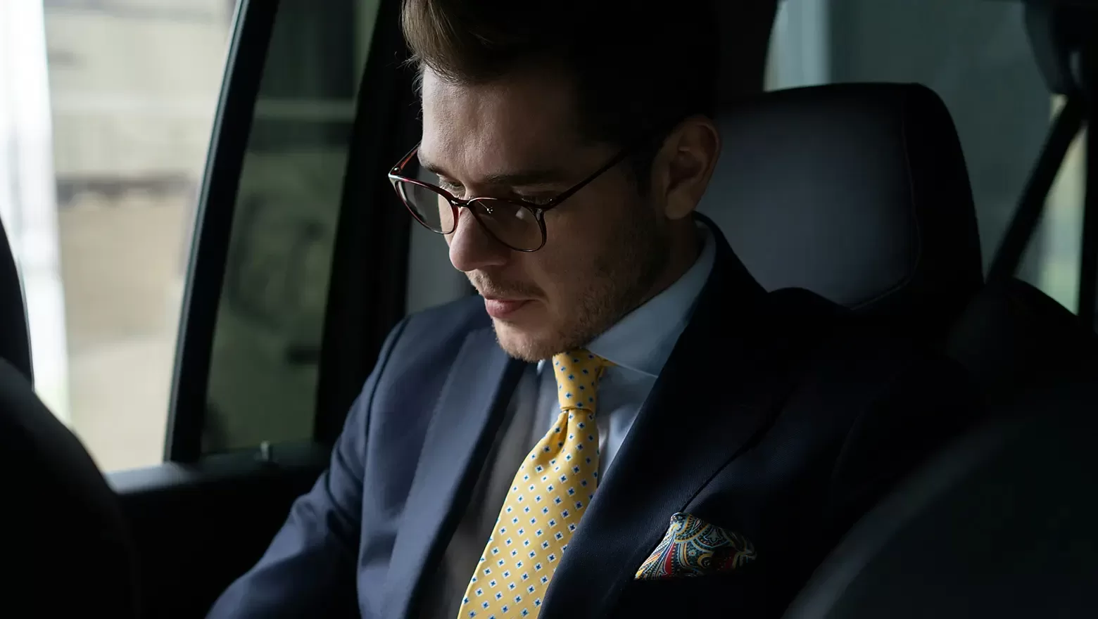 A man wearing glasses, a dark suit, a white shirt, and a yellow polka dot tie sits in the backseat of a car, looking down thoughtfully.