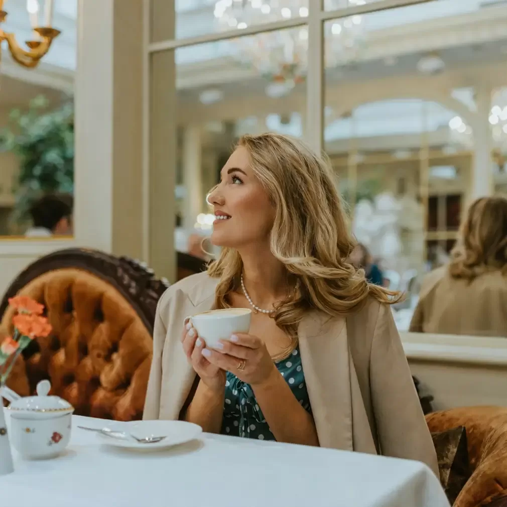 A woman with blonde hair sits at a table in an elegant café, holding a white teacup and smiling while looking upward. She wears a beige coat over a blue dress, and ornate mirrors and plush seating are visible in the background.