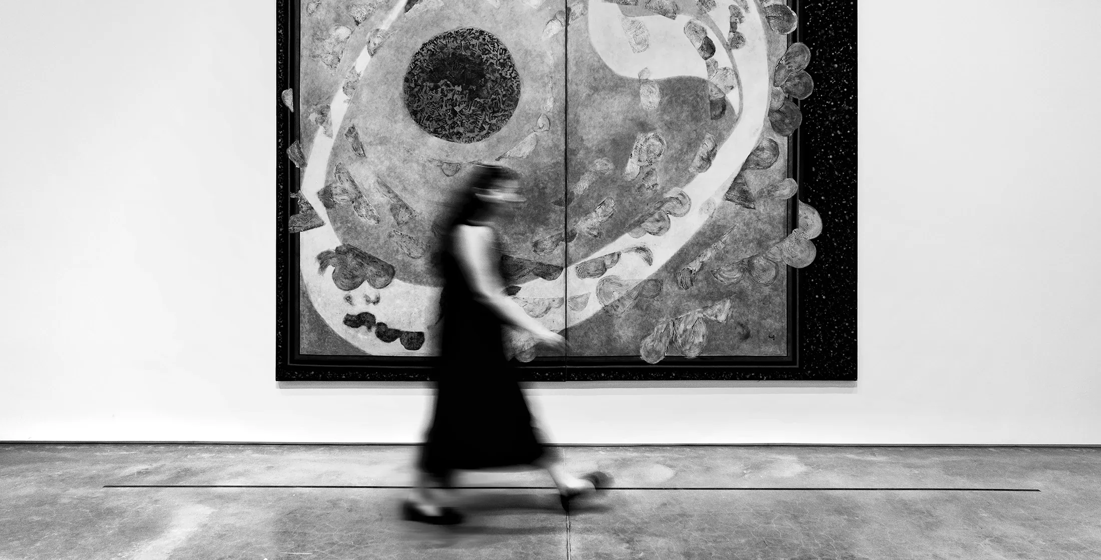 Black and white long exposure of a woman walking in front of a painting on a wall in a gallery.