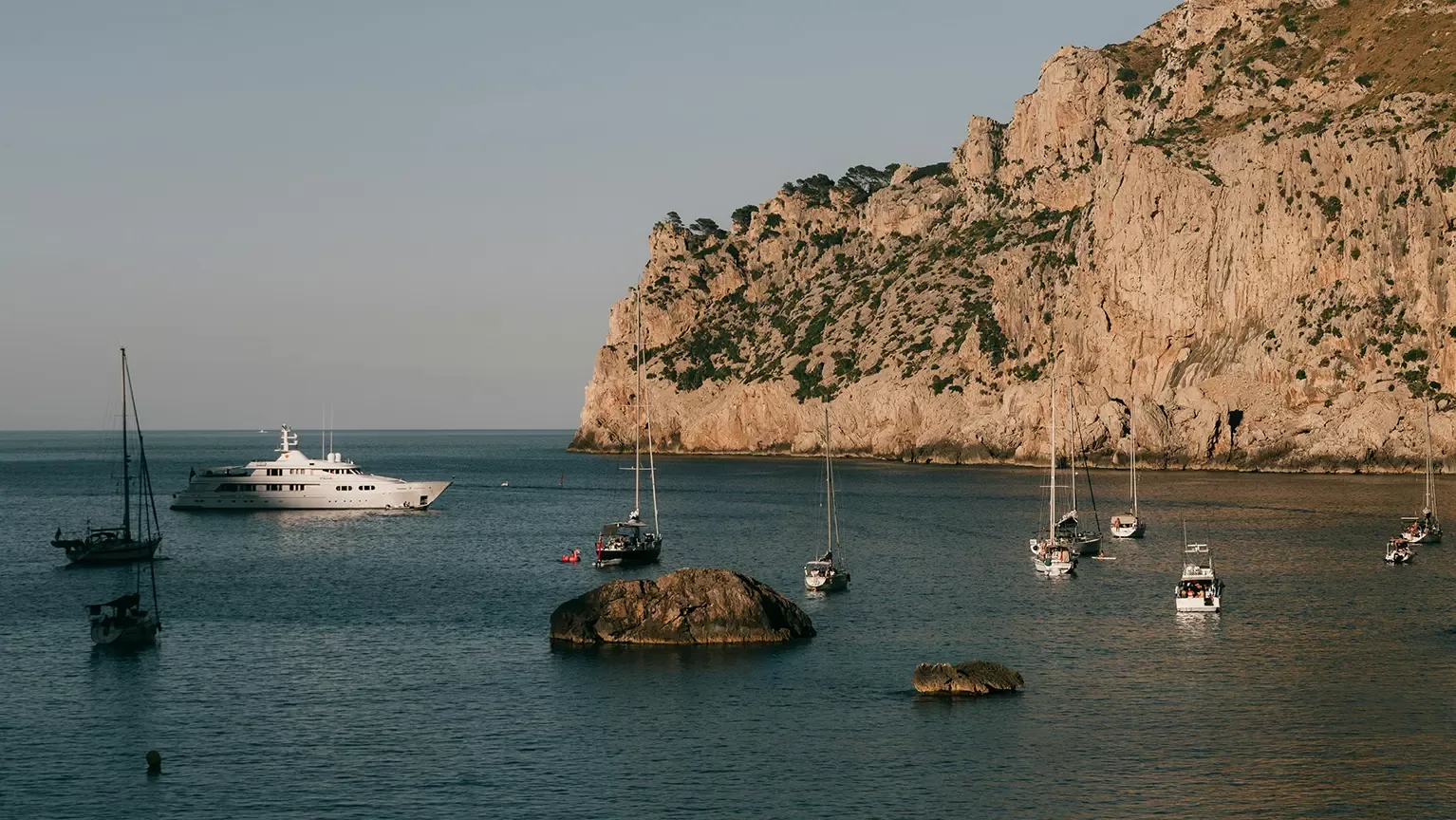 Several sailboats and a large white yacht are anchored in calm blue water near rocky cliffs, with scattered rocks in the bay and a clear sky above.