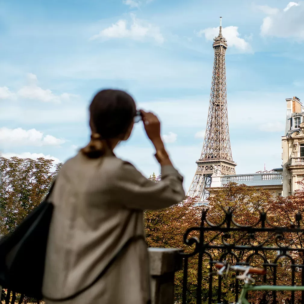 A person with a ponytail and a bag looks at the Eiffel Tower in Paris on a sunny day, with autumn-colored trees and city buildings in the background.