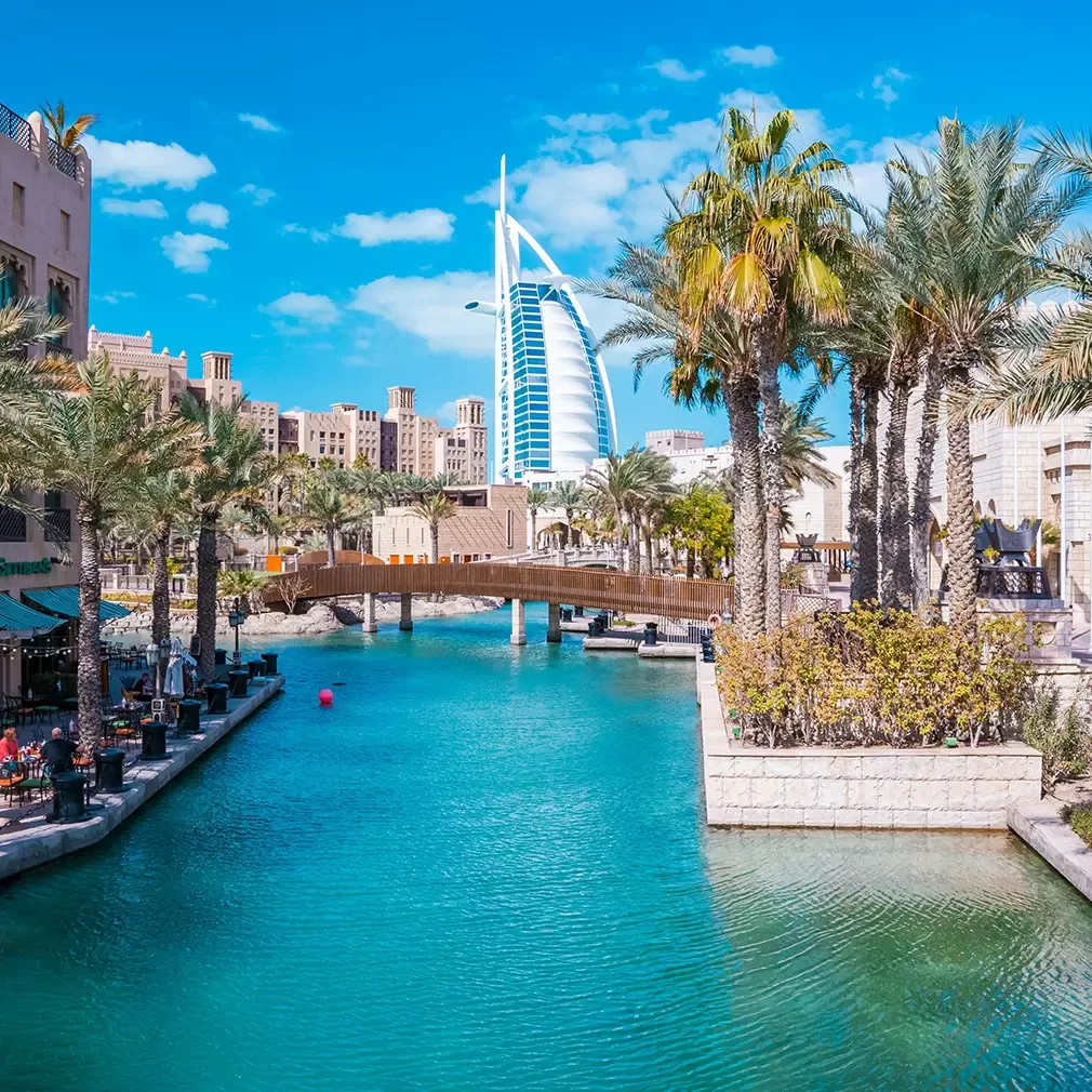 A canal lined with palm trees and tan buildings, with a wooden bridge crossing over it and the sail-shaped Burj Al Arab hotel visible in the background under a bright blue sky.
