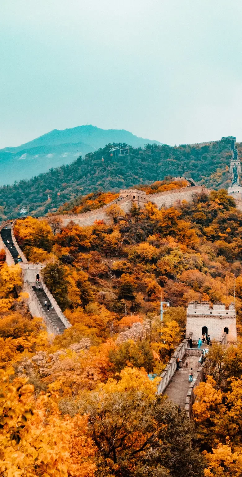 The Great Wall of China winding through lush, autumn-colored hills, with vibrant orange and yellow foliage. Towering mountains are visible in the background under a cloudy blue sky.