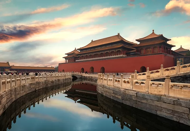 The Forbidden City in Beijing, China, at sunset. Traditional red and gold palace buildings are reflected in a curved moat, with ornate stone railings and a dramatic sky above.
