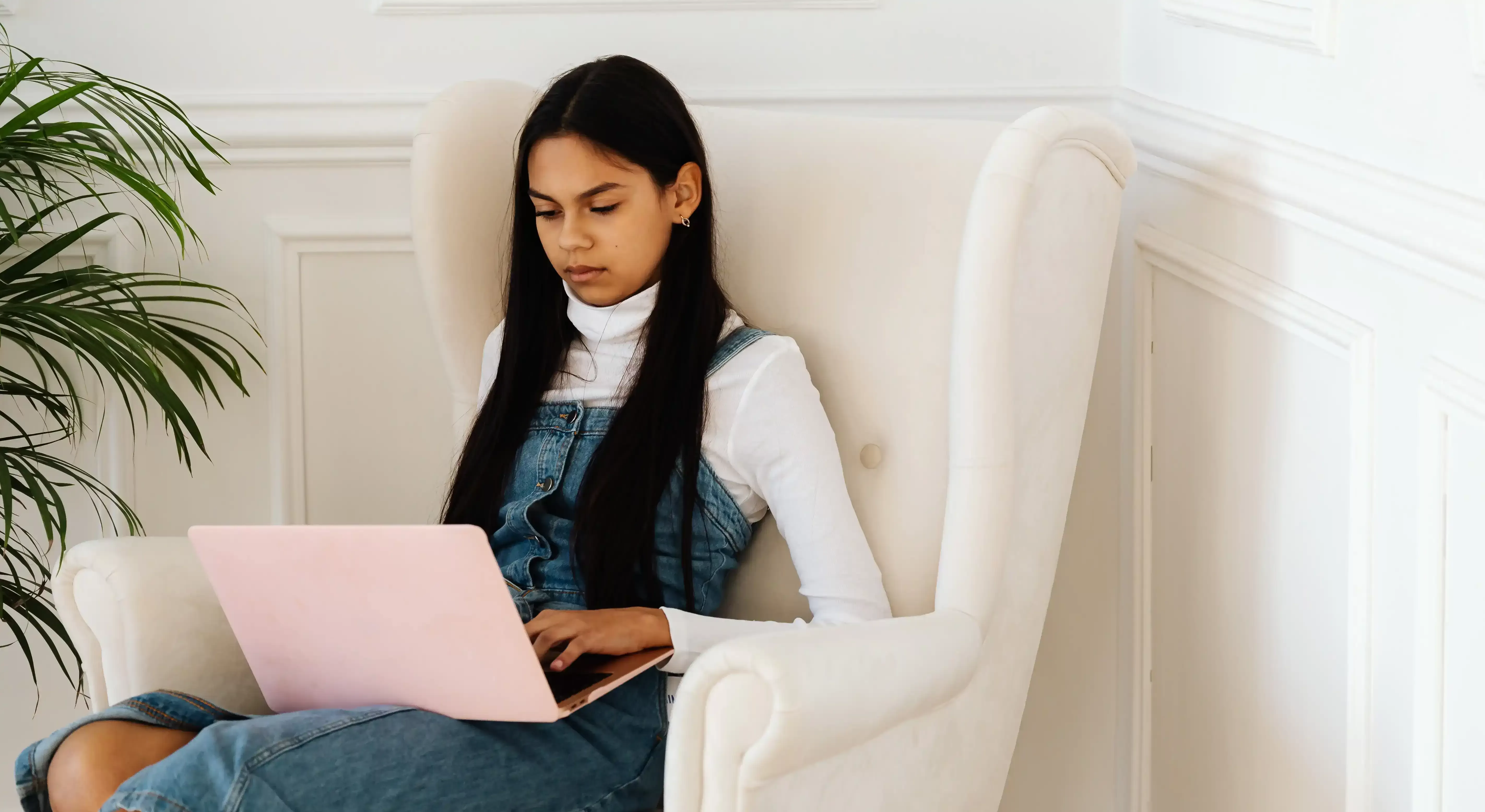 Women with a pink laptop on her lap sat on a white arm chair next to a plant