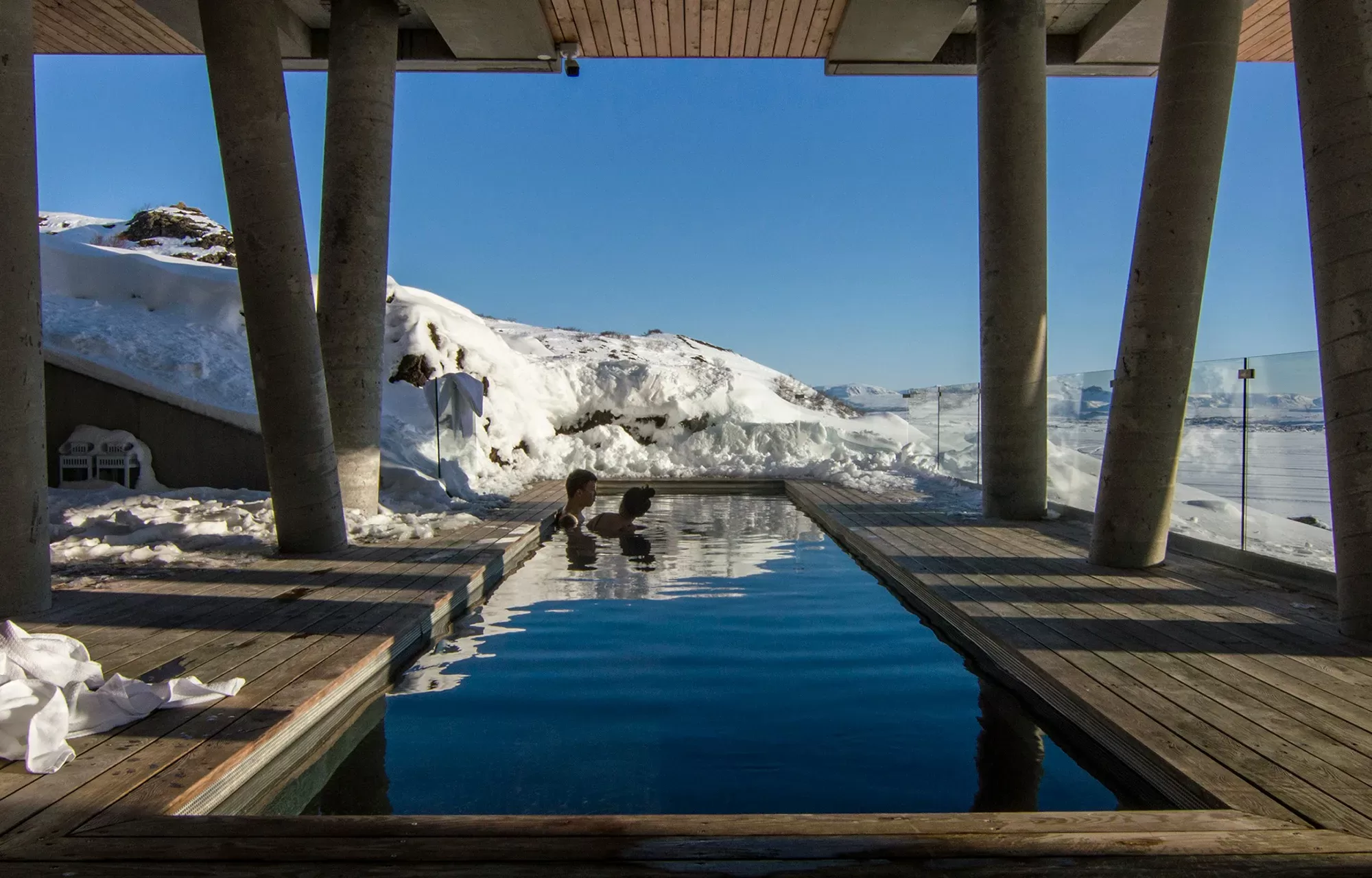 Two people enjoying a warm outdoor pool surrounded by snow, with a view of snow-covered mountains under a clear blue sky.