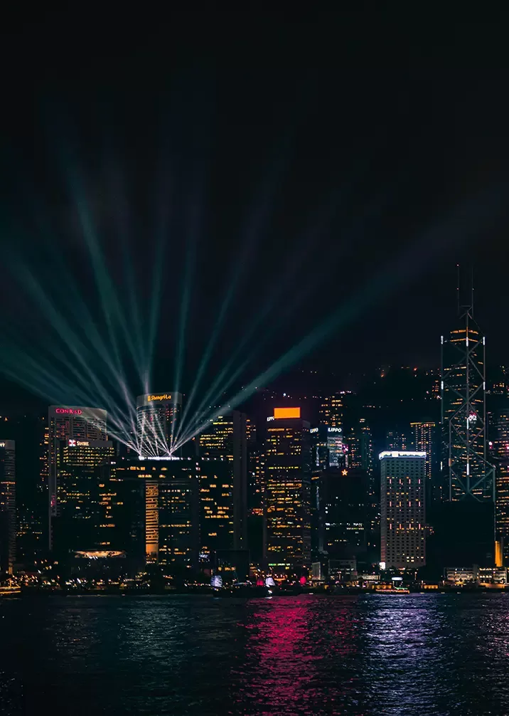 A nighttime cityscape featuring a skyline with illuminated skyscrapers. Bright lights from buildings and laser beams create a vivid display, reflecting on the water below, adding to the scenes vibrant atmosphere.