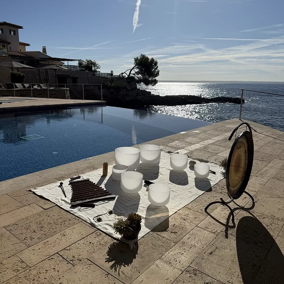 A set of white crystal singing bowls, mallets, a gong, and other sound healing instruments are arranged on a white cloth beside an infinity pool overlooking the sea on a sunny day.