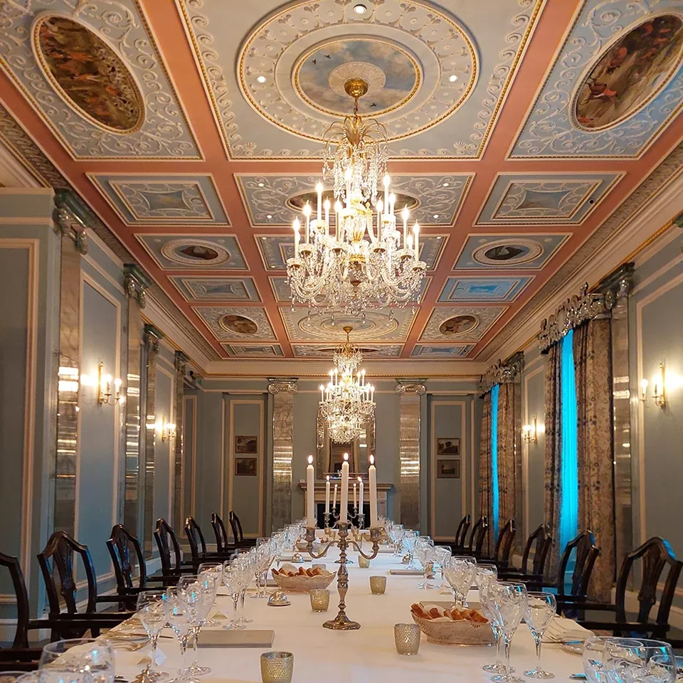 An elegant dining room with ornate chandeliers, detailed ceiling art, and long table set with candles, glasses, and plates. The room features tall windows, pale blue walls, and decorative columns.