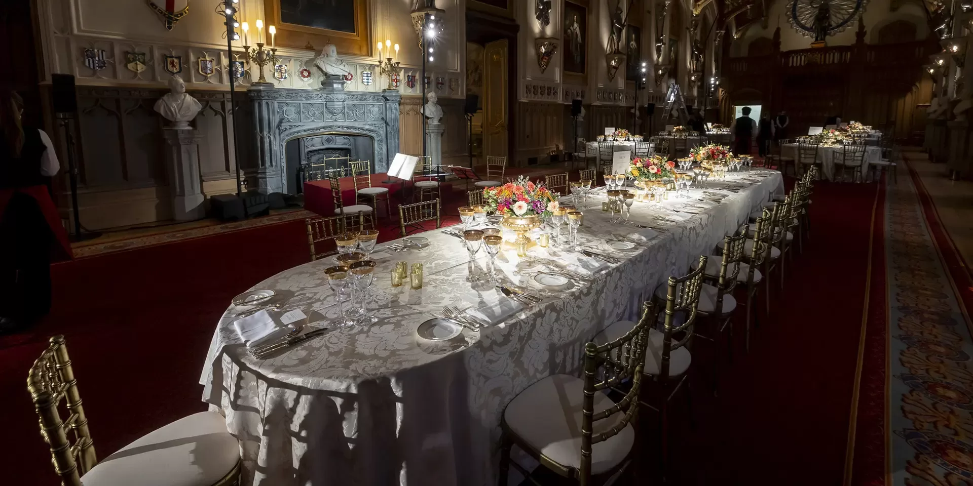 A long, elegant dining table with white lace tablecloth, set with fine china, glassware, and floral centerpieces in a grand hall with ornate décor, chandeliers, and a large decorative fireplace.