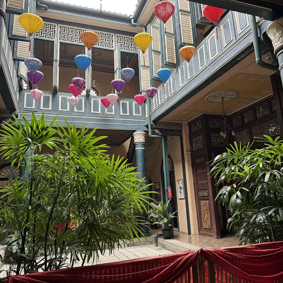 A courtyard with tropical plants and colorful lanterns hanging above. The walls feature ornate designs and wooden balconies, giving the space a traditional, historic look. Red drapes are in the foreground.