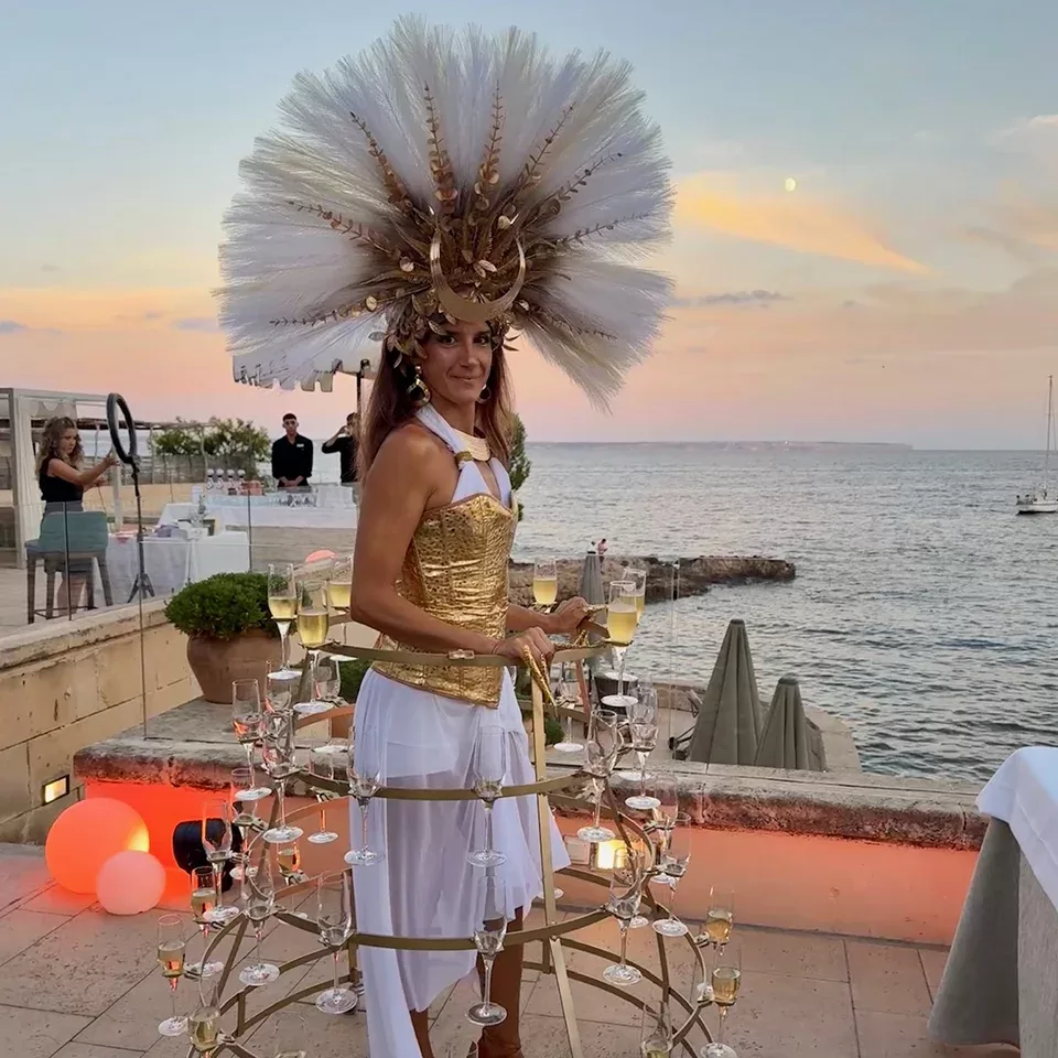 A woman in a gold corset and elaborate headdress stands by the sea at sunset, wearing a hoop skirt that holds multiple glasses of champagne. The scene is festive with elegant outdoor decor and a soft pastel sky.