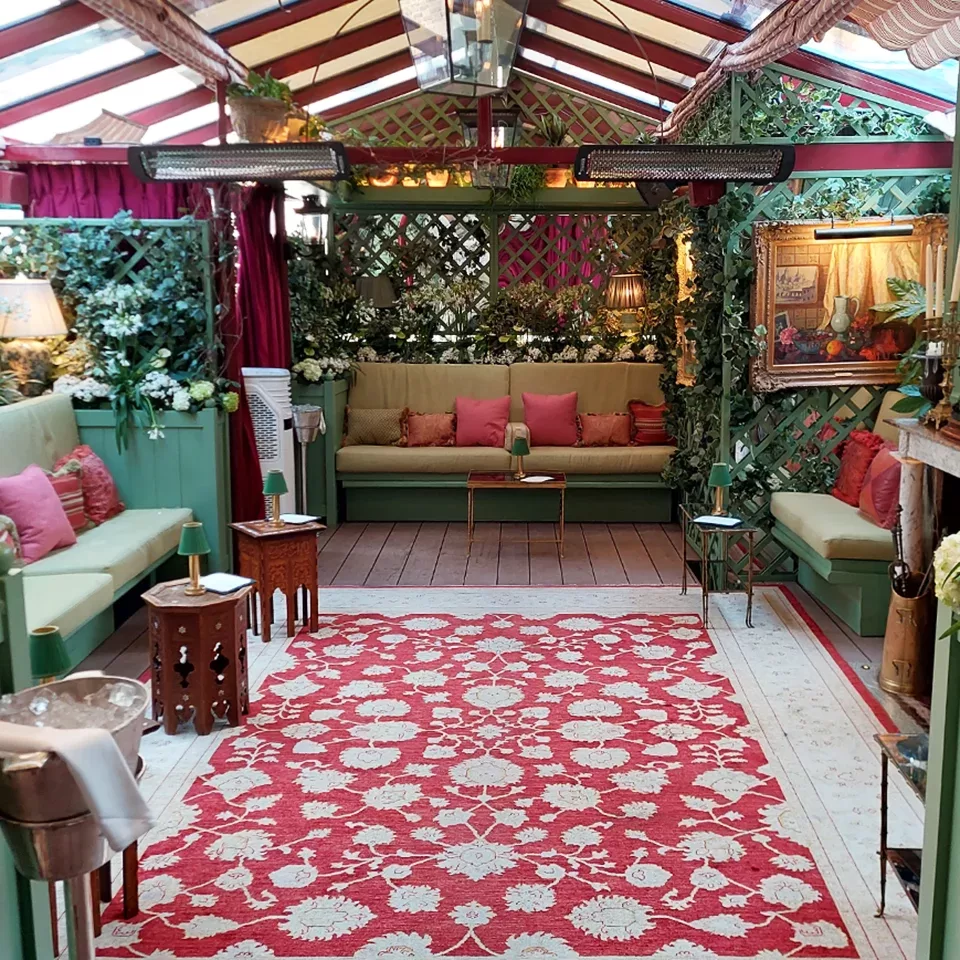 A cozy indoor lounge with green walls, cushioned benches with pink pillows, a red and white floral rug, plants, soft lighting, and paintings creating an inviting, garden-inspired atmosphere.