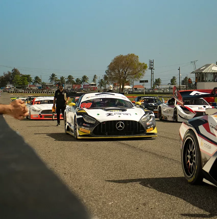 Several race cars, including a prominent white Mercedes-Benz with the number 88, line up on a racetrack under a clear sky. A person and other cars are visible, with palm trees in the background.