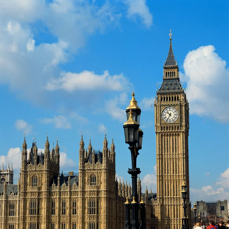 The photo shows the iconic Big Ben clock tower and part of the Houses of Parliament in London under a bright blue sky with scattered clouds. A decorative street lamp is visible in the foreground.