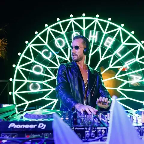 Dj wearing sunglasses playing on desks in front of lit up 'Coachella' sign