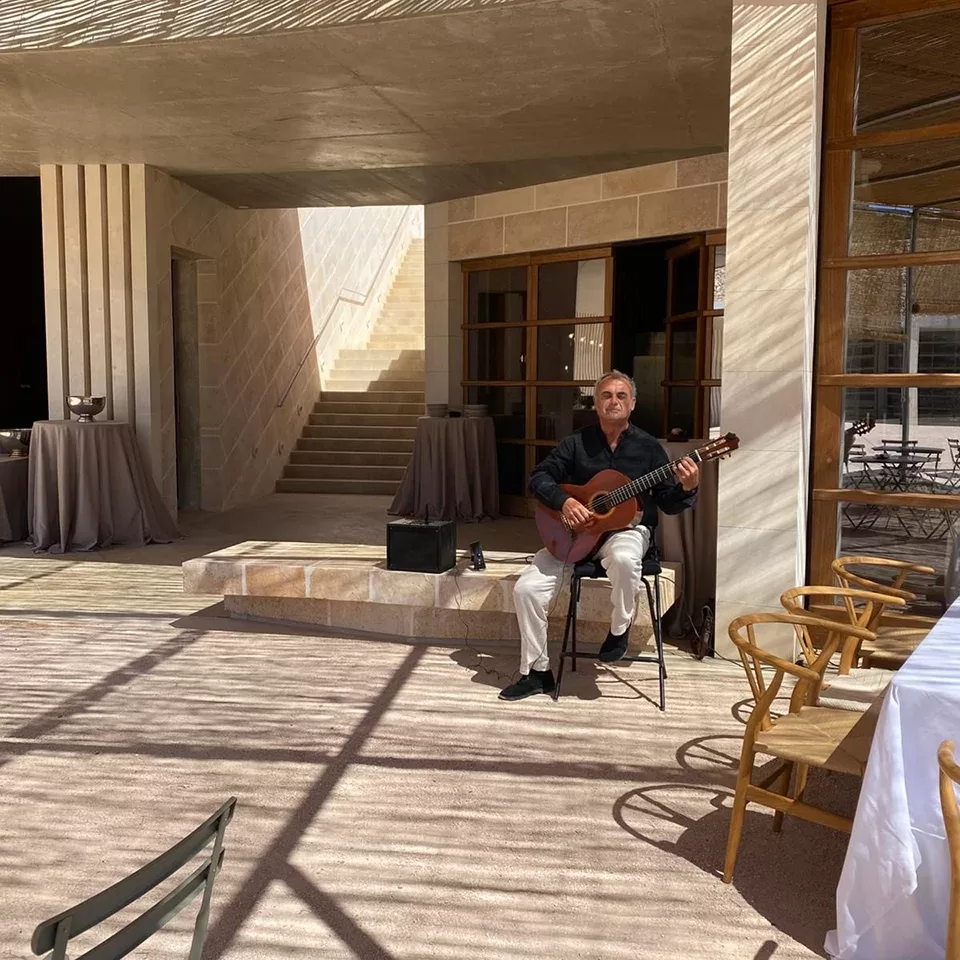 A man sits on a stool playing a classical guitar in a sunlit outdoor area with stone walls and tables covered in gray cloth; wooden chairs and shadows from above are visible.