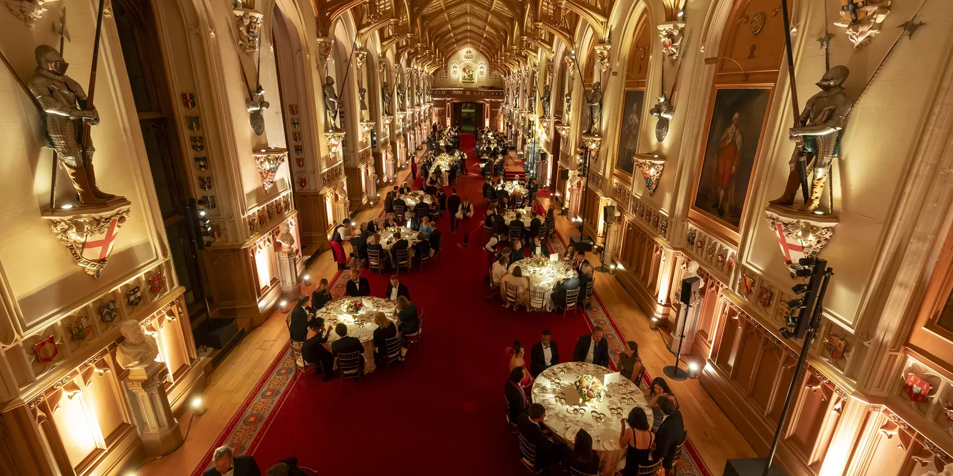 A grand, ornate hall with high vaulted ceilings and statues, filled with round tables where people are dining, all illuminated by warm, golden lighting.