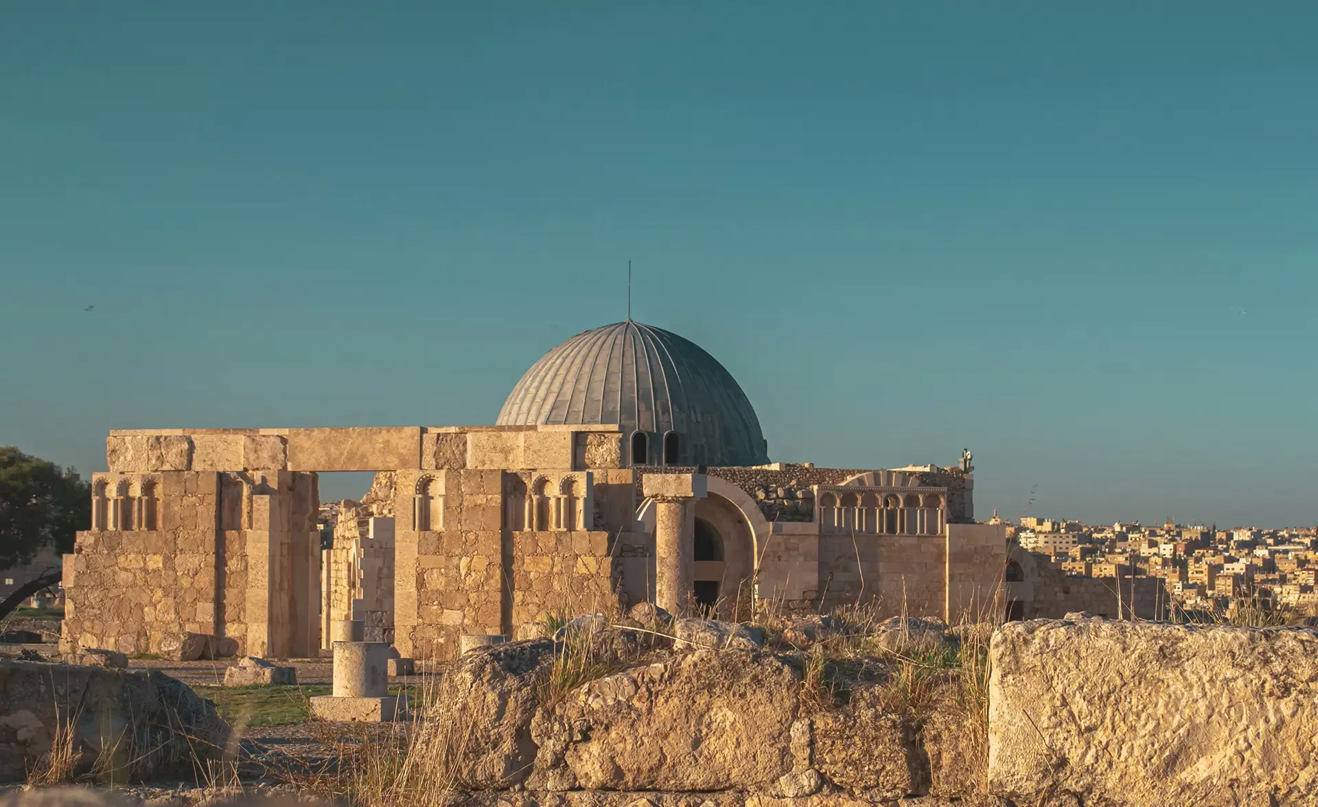 A historic stone building with a large central dome under a clear blue sky, surrounded by ancient ruins. Part of a sprawling cityscape is visible in the background.