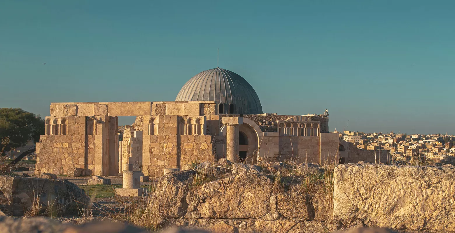 A historical stone structure with a prominent domed roof stands under a clear blue sky. The ancient architecture is surrounded by rocky terrain, with a cityscape visible in the background.