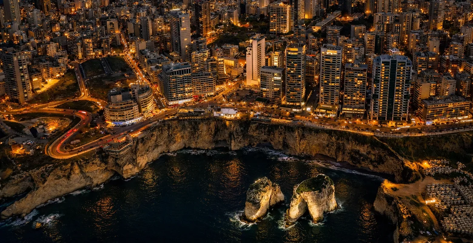 Aerial view of a coastal city at night, with illuminated high-rise buildings lining cliffs above the sea. Two large rock formations rise from the dark water near the shore. Streets are lit with yellow lights, creating a vibrant cityscape.