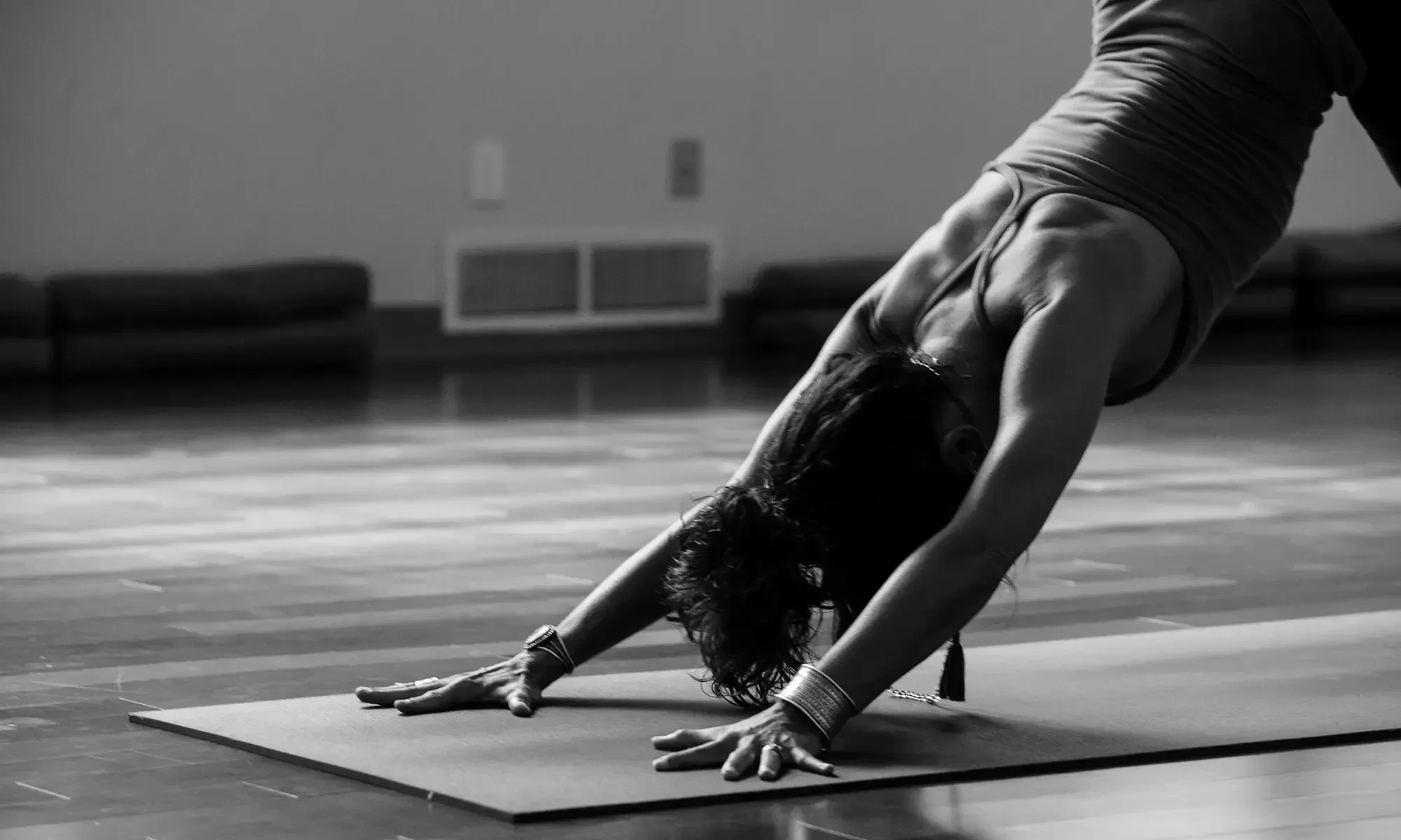 A person performs the downward dog yoga pose on a mat in a dimly lit room. They are wearing a tank top and bracelets, with their head down and arms extended forward. The floor is wooden, and cushions are visible in the background.