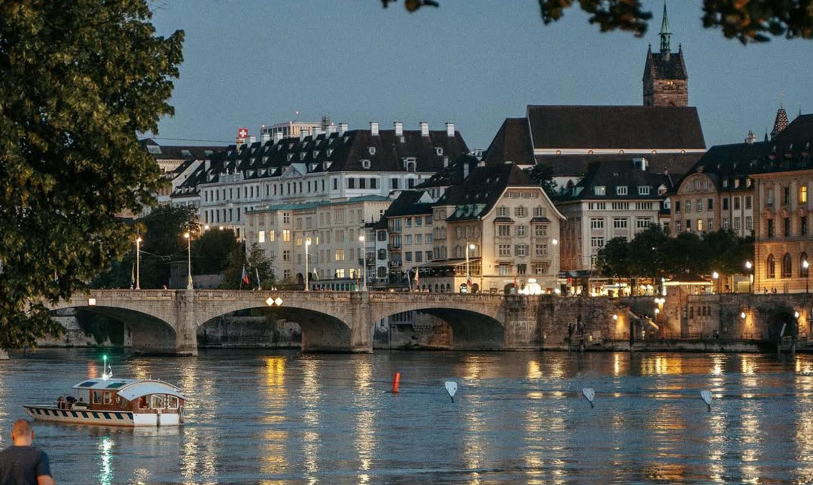 A scenic evening view of a European city riverfront with historic buildings, a church with a tall spire, a stone bridge, and a small boat on the calm water, framed by leafy trees.