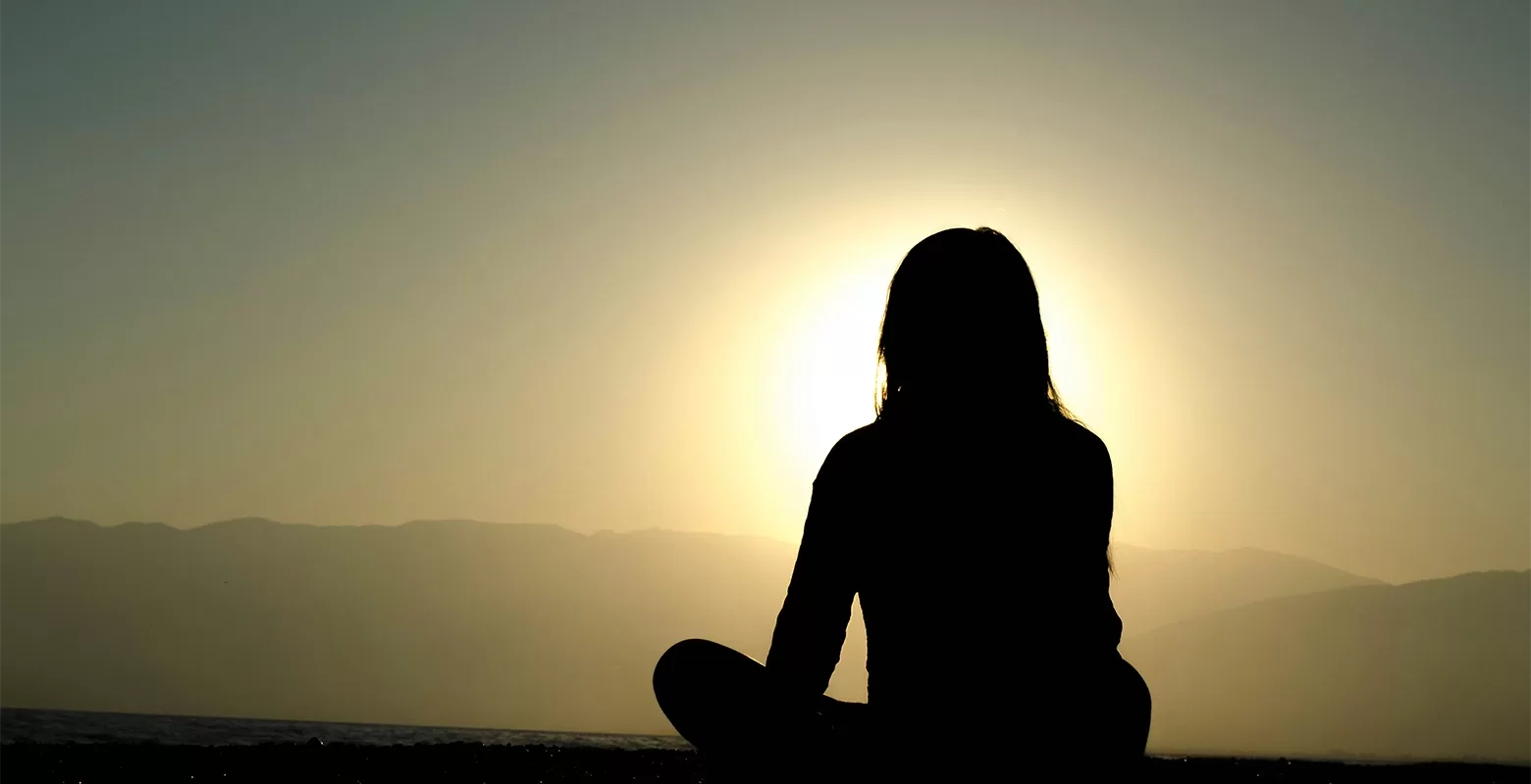 Silhouette of a woman doing yoga on the floor cross legged facing the mountains and the sunset.