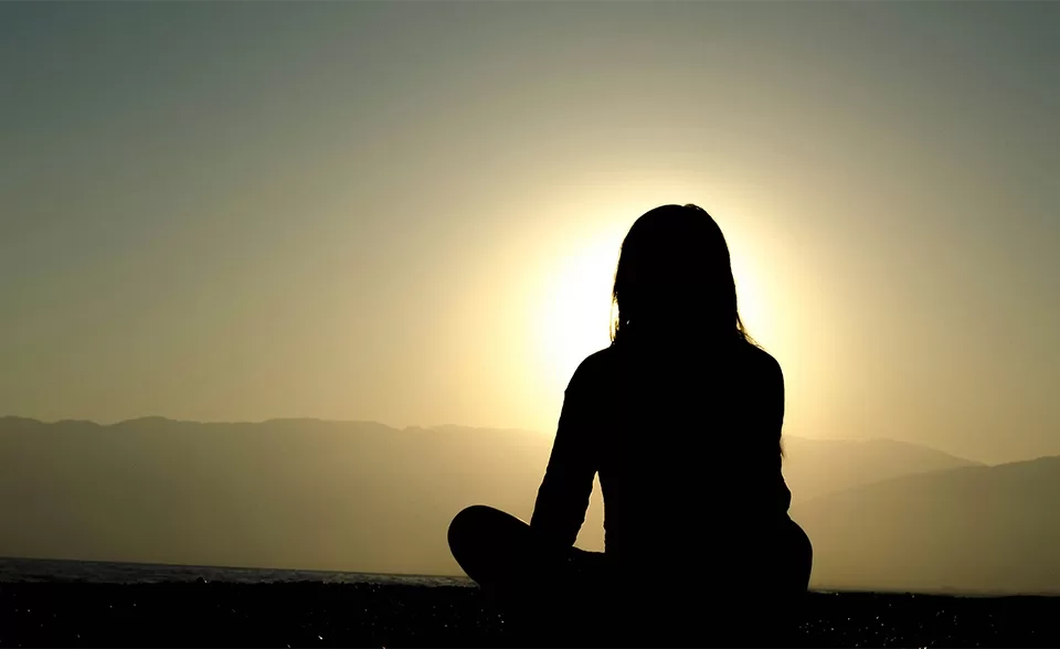 Silhouette of a woman doing yoga on the floor cross legged facing the mountains and the sunset.