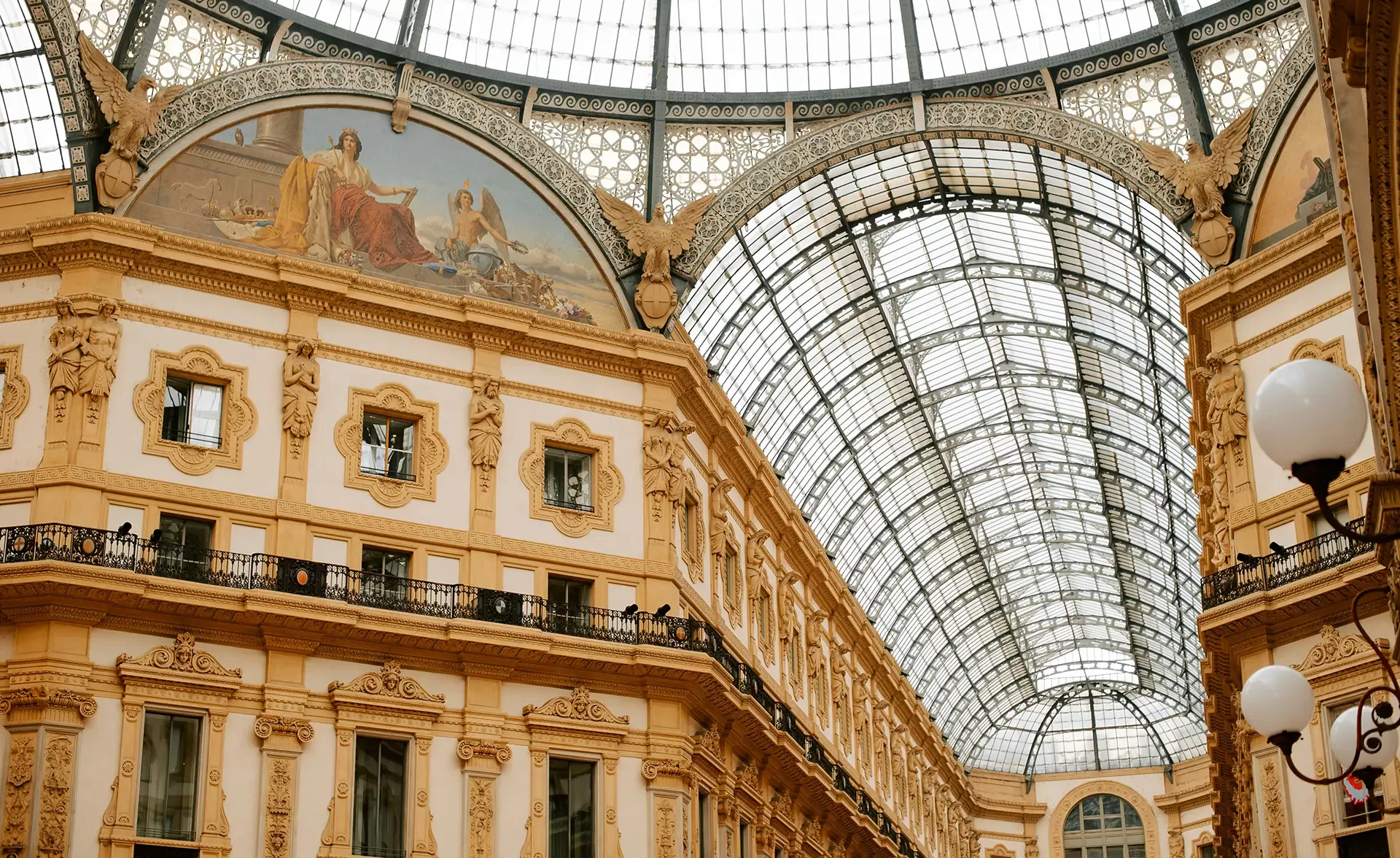 Interior of a grand gallery with a high, arched glass ceiling and ornate architectural details. The walls are decorated with intricate designs and a large fresco. A streetlamp is visible in the foreground.