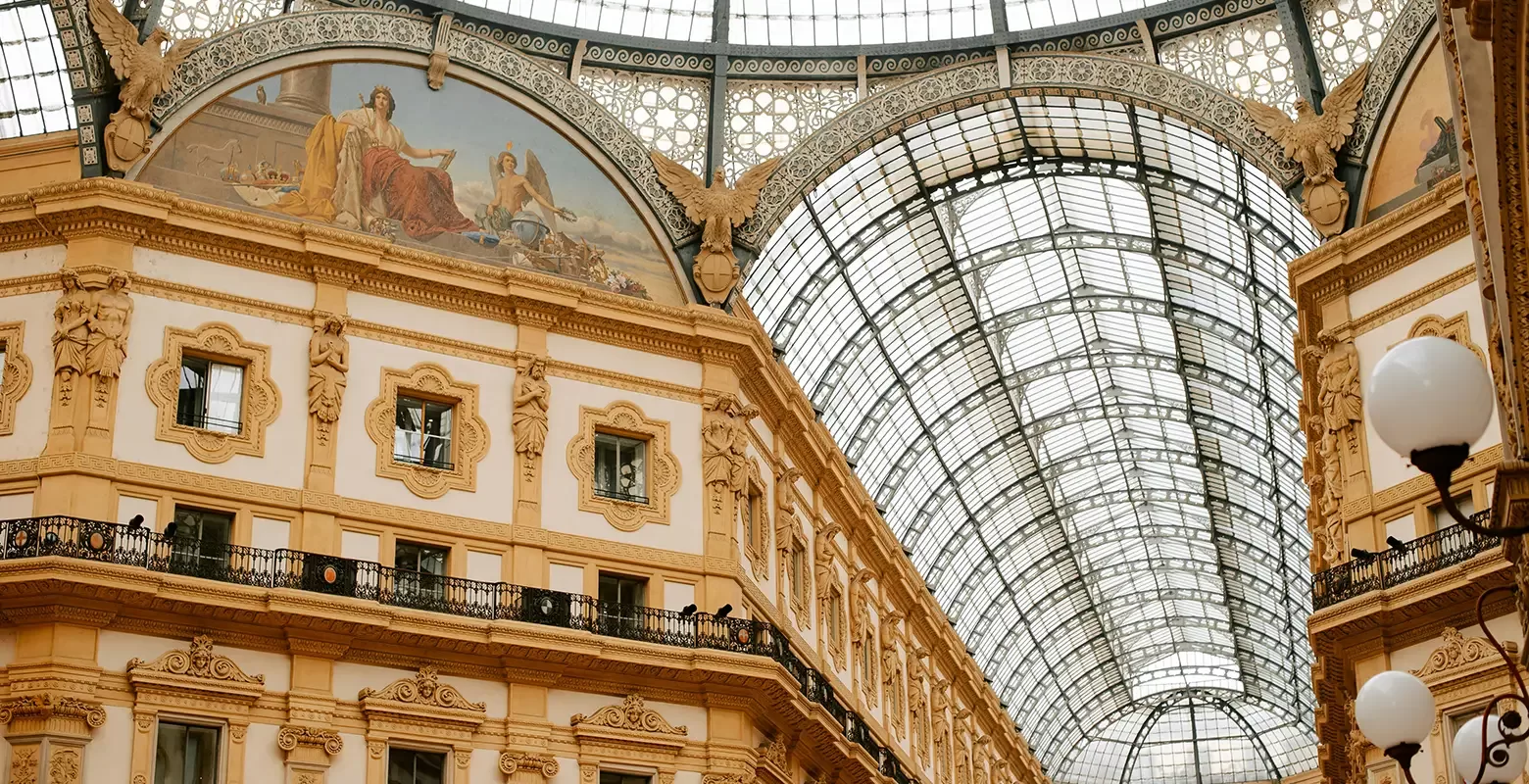 Interior view of a historic shopping gallery with a curved glass roof and ornate architectural details. The building features intricate moldings, a fresco on the upper wall, and decorative arches, bathed in natural light.