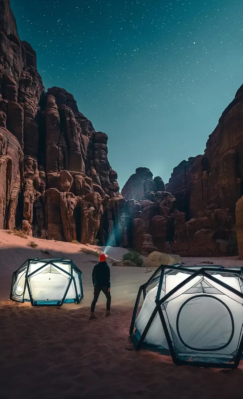 Man wearing head torch looking up at starry sky next to two lit up tents in the desert of Neom, Saudi Arabia