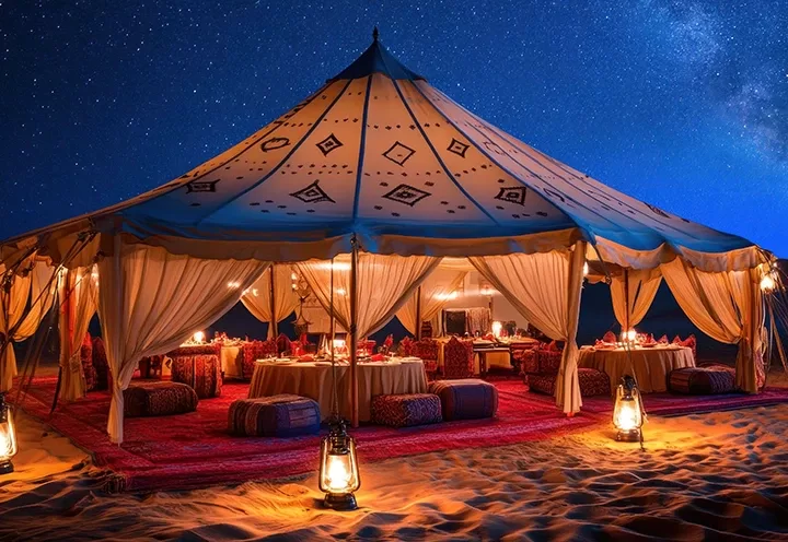 A luxurious desert tent decorated with lights and lanterns sits on sand under a starry night sky, with low tables and colorful cushions inside, creating a cozy, inviting atmosphere.