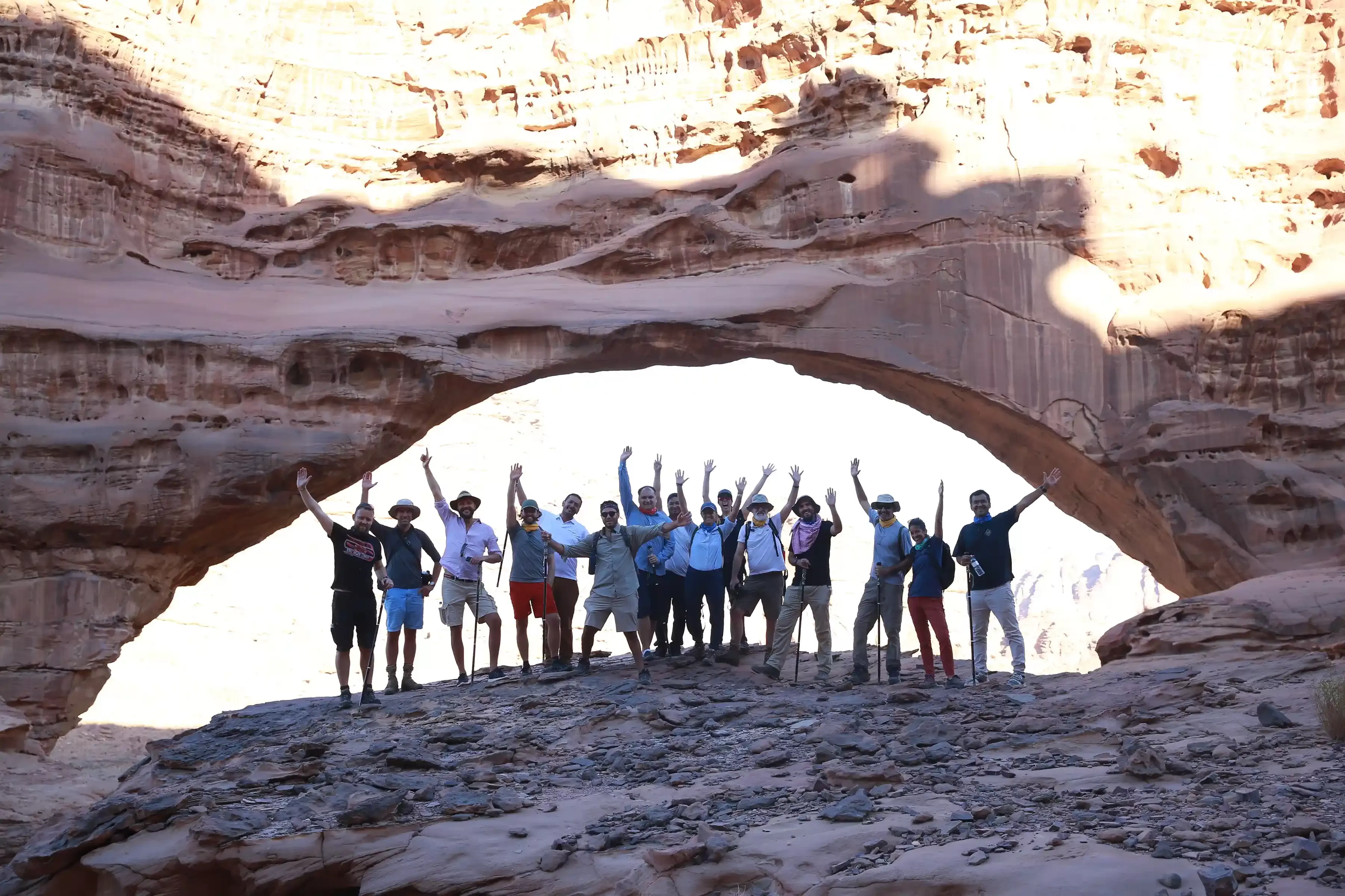 Quintessentially | Group of people in summer clothes and their hands in the air stood under a large arched rock in Neom, Saudi Arabia
