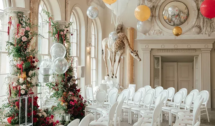 A bright, ornate room set up for an event, with rows of white chairs facing towards a floral archway. Colourful balloons, a giraffe statue mounted on the wall, large windows, and a decorated ceiling add whimsical details to the elegant space.