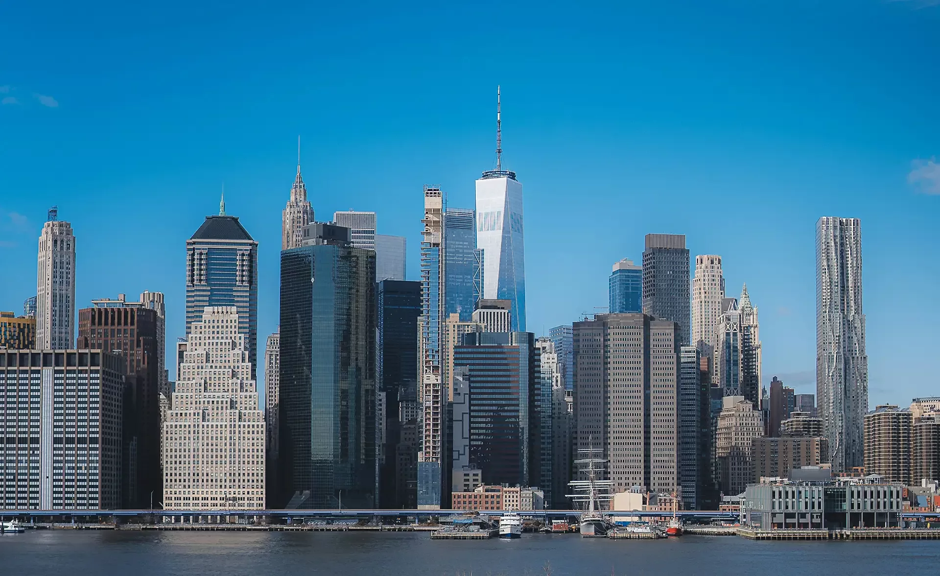 A panoramic view of a citys skyline featuring tall, modern skyscrapers under a clear blue sky. The buildings are reflected in the calm water in the foreground, creating a symmetrical visual effect.