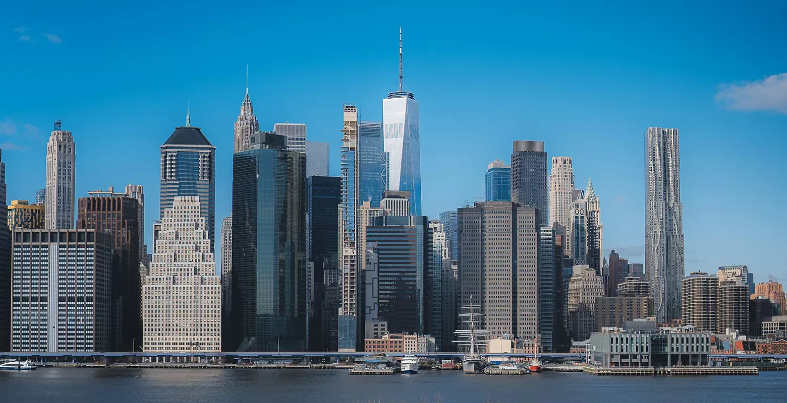 A panoramic view of a city skyline featuring tall, modern skyscrapers under a clear blue sky. The buildings reflect sunlight, and the waterfront is visible in the foreground.